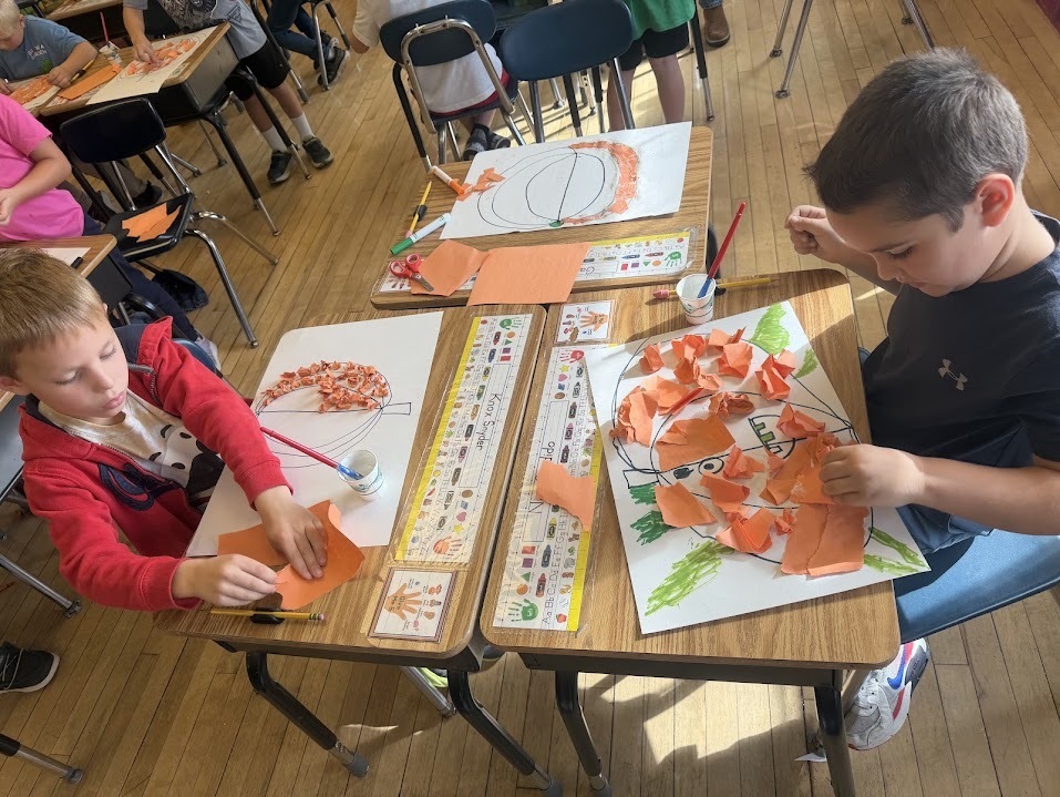 Two students sitting side by side working intently on gluing pieces of orange construction paper inside large pumpkin drawings on paper.