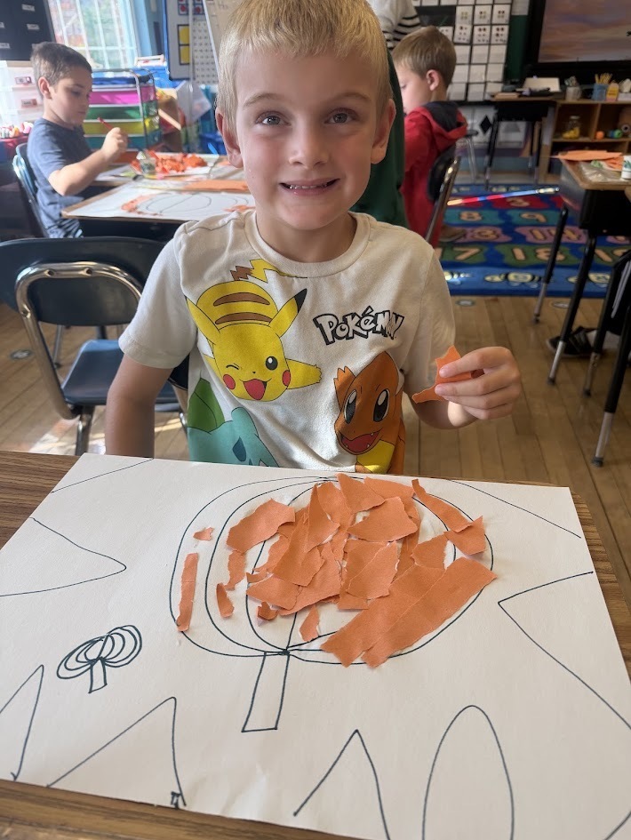 A smiling boy wearing a Pokémon t-shirt holding a strip of orange paper over his finished paper pumpkin collage.