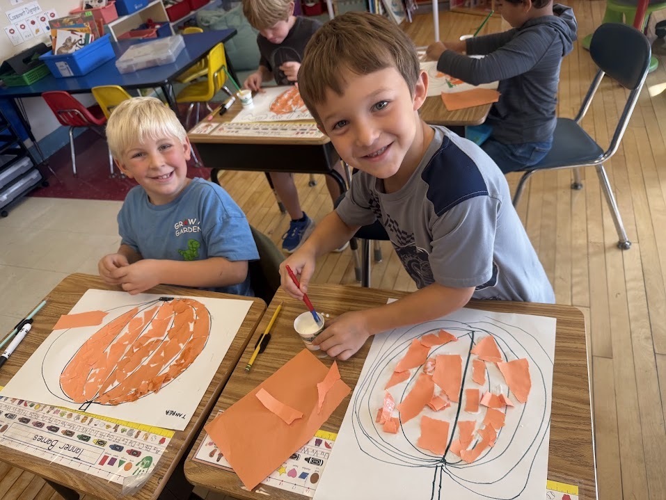Two young boys smiling at the camera as they use small paintbrushes to apply liquid glue onto their pumpkin art projects.