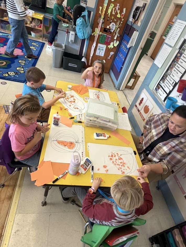 An overhead shot of a classroom activity table where four young students and an educator sit together working on paper pumpkin crafts.