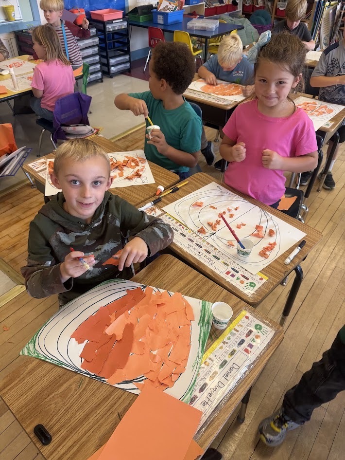 A group of young elementary students sitting at their classroom desks, happily pasting torn orange paper onto pumpkin outlines for a fall art craft.