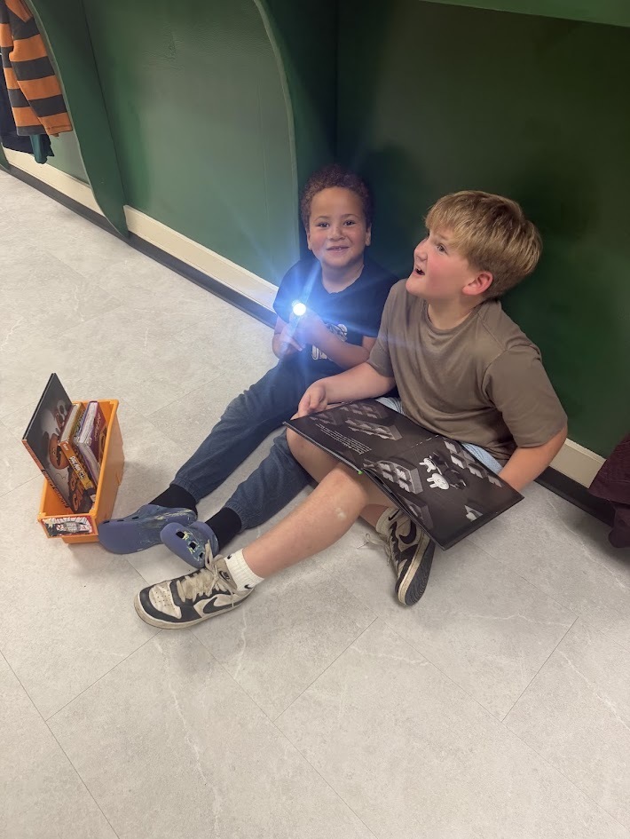 Two young boys sitting on the floor in a hallway reading an open book with a flashlight.