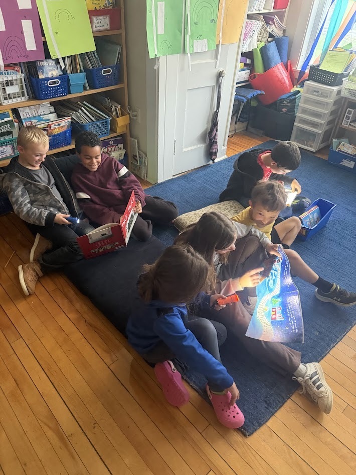 A group of elementary students sitting cross-legged on a classroom rug, using small flashlights to read books together.
