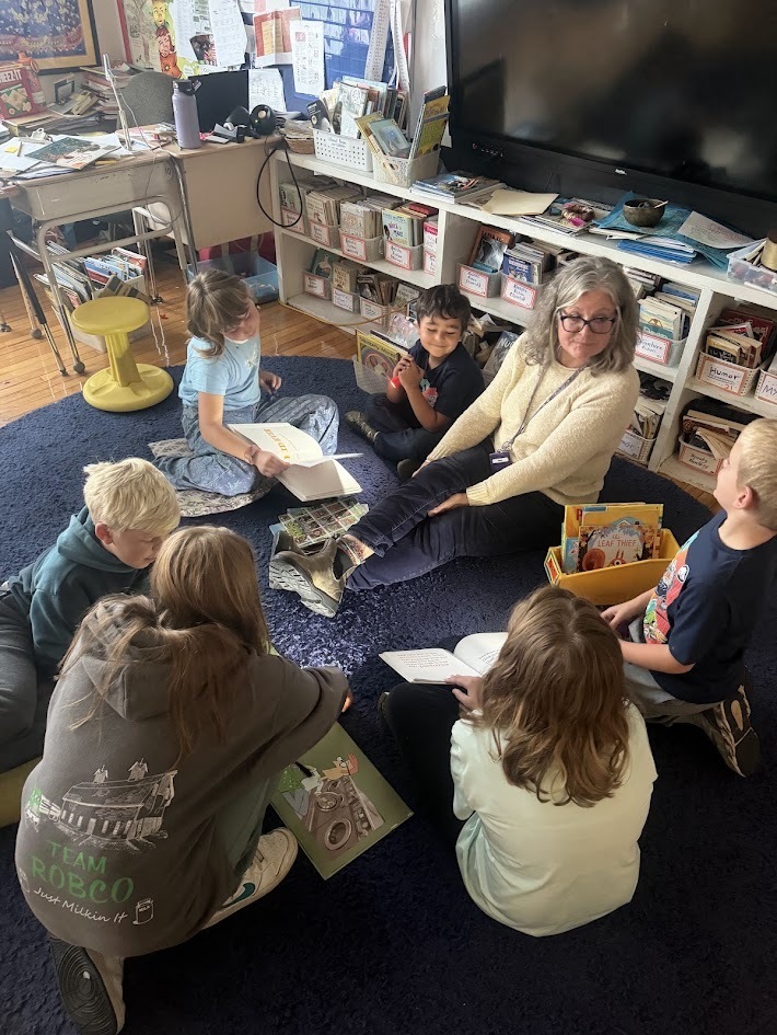 A teacher sitting in a circle on a large blue rug with a group of elementary students as they hold flashlights and read their books.