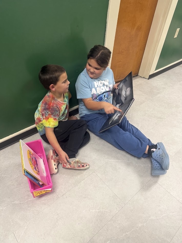 Two students sitting together on the hallway floor leaning against a wall while reading a picture book together.