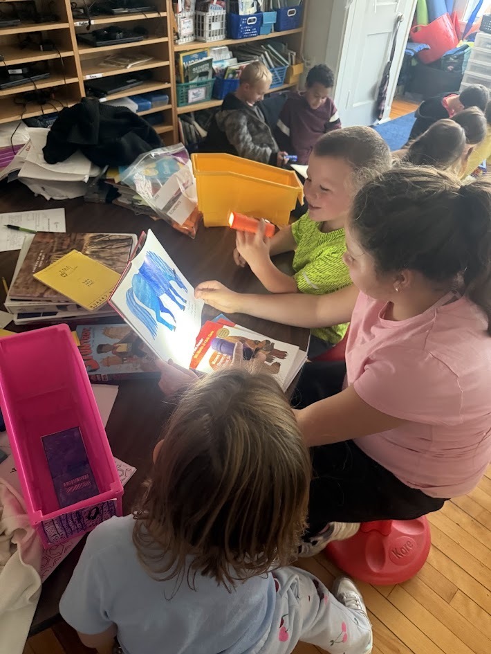 Three students sitting on flexible stools around a small table, using a flashlight to shine light on a picture book as they read together.