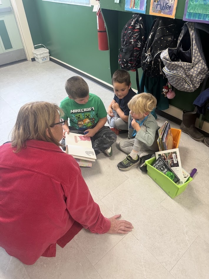 An adult seated on the hallway floor listening as three young students share and read a book together.