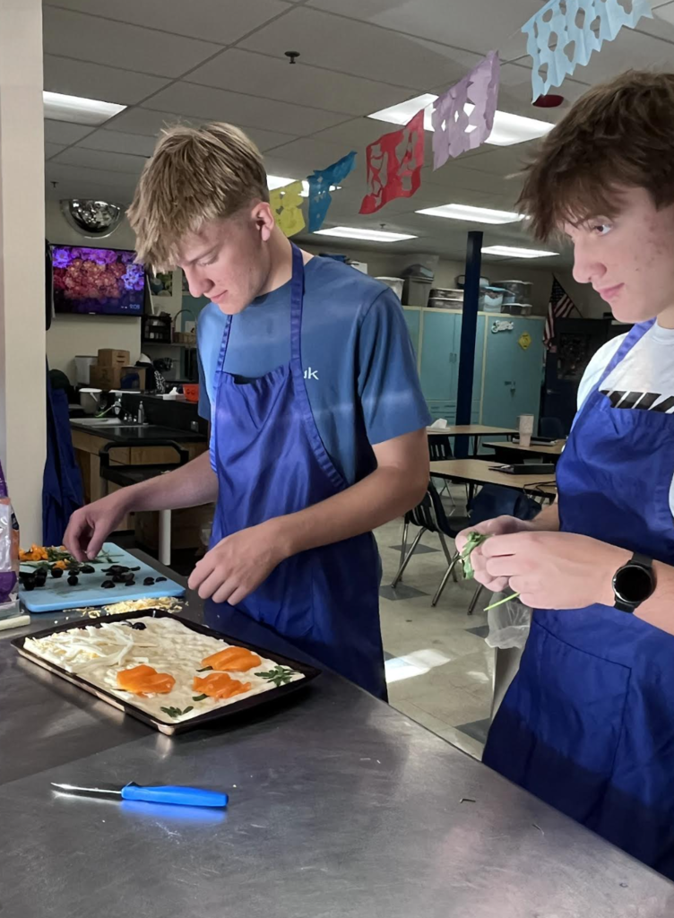 Two male students wearing blue aprons working together at a stainless steel kitchen counter, carefully arranging sliced peppers, herbs, and olives on a sheet of focaccia dough to create a decorative floral artwork.