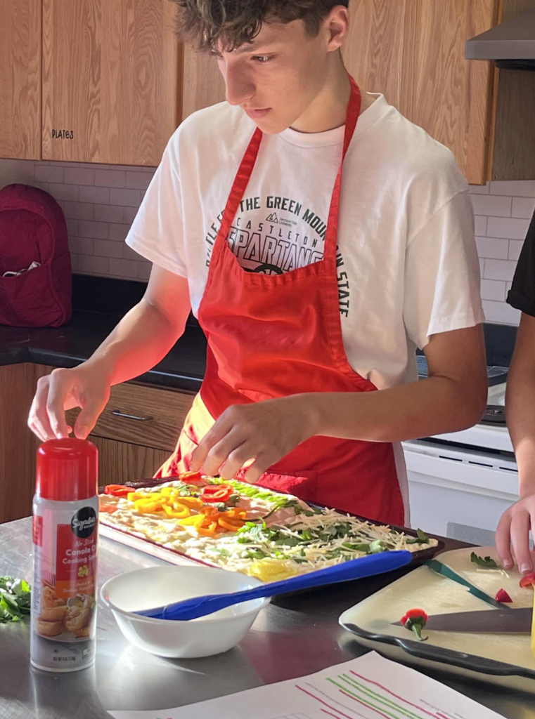 A male student wearing a red apron over a white Castleton Spartans shirt carefully arranging bright yellow and red sliced peppers onto a tray of raw focaccia dough.