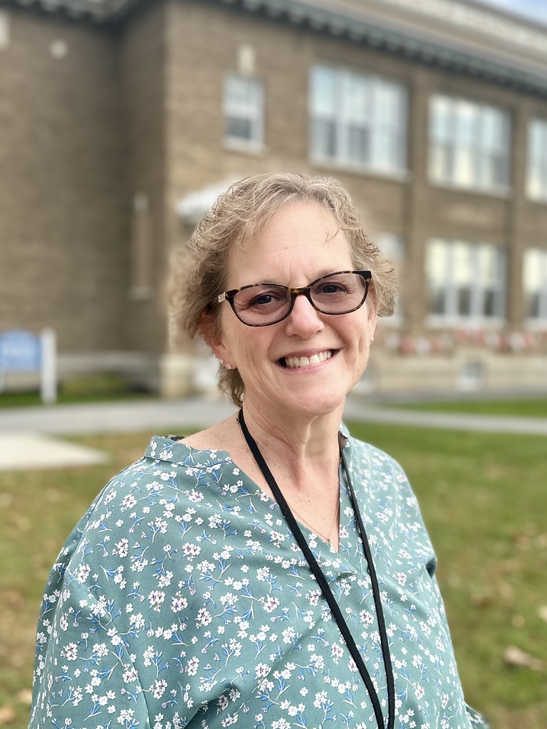 Photo of next year's Principal at Fair Haven Grade School, she is standing in front of the school, smiling, wearing a green floral shirt