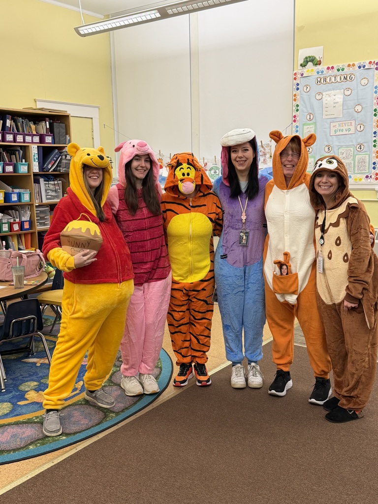 Six elementary school teachers dressed in colorful full-body animal onesies representing characters from Winnie the Pooh, including Winnie the Pooh holding a plush "HUNNY" pot, Piglet, Tigger, Eeyore, Kanga, and Owl. They are standing together inside a decorated classroom, smiling for the camera.