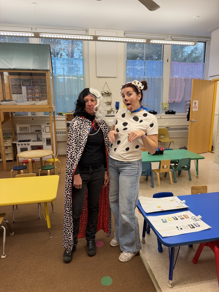 Two preschool teachers posing in a classroom dressed as 101 Dalmatians characters. One teacher is dressed as Cruella de Vil with a black and white wig, pearl necklace, and spotted cape. Beside her, another teacher is dressed as a Dalmatian puppy wearing a black-and-white spotted shirt, dog ears headband, and face paint, posing playfully.