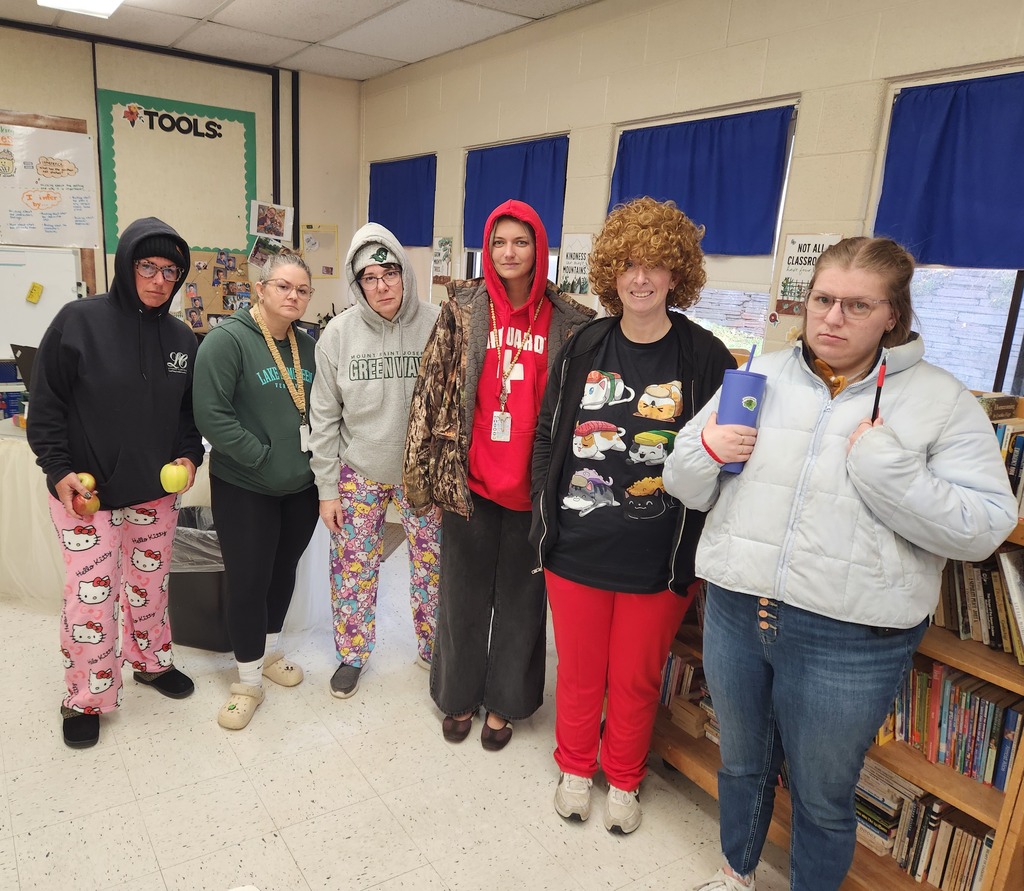 Six female teachers dressed as students standing together in a classroom, wearing hoodies, graphic pajama pants, Crocs, and a curly wig while holding apples and travel mugs.