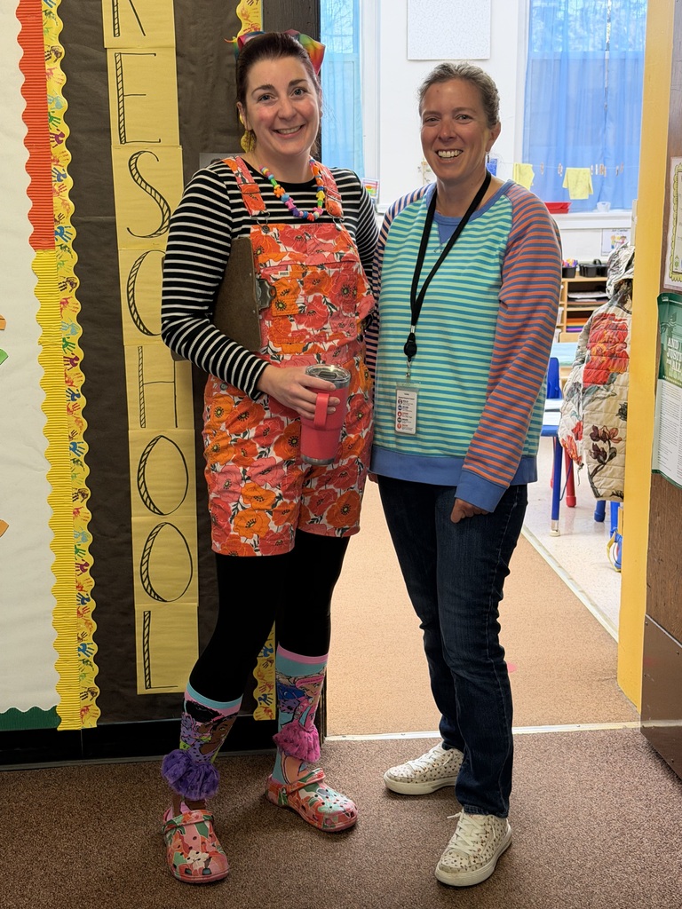 Two teachers dressed in bright, playful outfits posing in a school doorway, with one wearing floral overalls and colorful socks while holding a clipboard and travel tumbler.