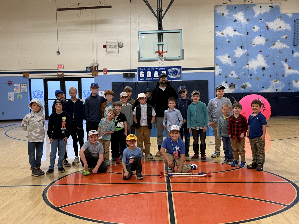 A large group of students dressed as teachers posing on the school gym basketball court, wearing button-down shirts, slacks, baseball caps, fake facial hair, and school lanyards.