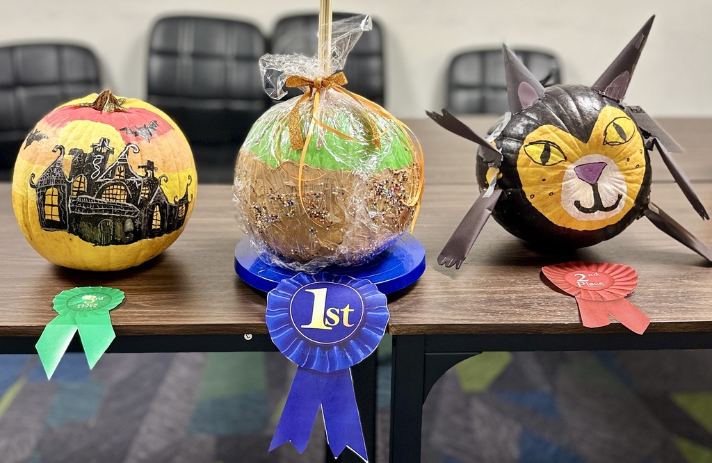 The top three winning contest pumpkins lined up on a table with award ribbons: 3rd Place painted haunted house, 1st Place wrapped caramel apple, and 2nd Place black cat.