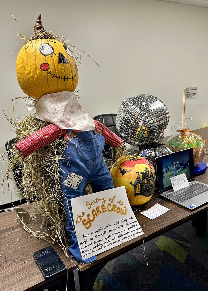 A pumpkin decorated as a full scarecrow in overalls named "Sorting of the Scarecrow," sitting on a table next to a disco ball pumpkin and a haunted house pumpkin.