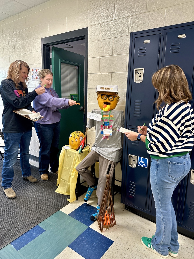 Principal Paquette, Ms. Ezzo, and Ms. Pelletier holding clipboards while judging a life-sized pumpkin figure dressed in a t-shirt and hat by school lockers.