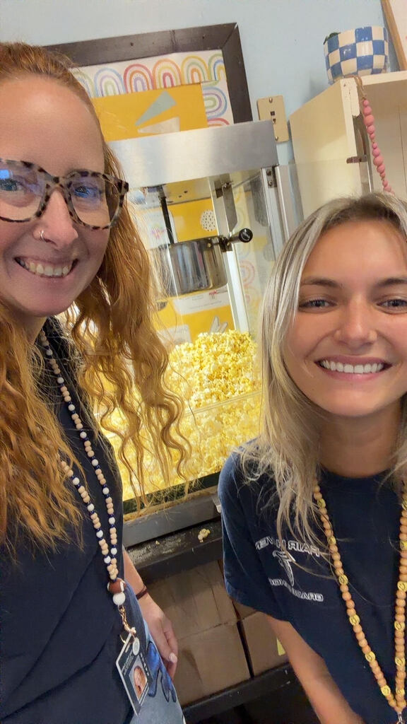 Two staff members smiling for a selfie in front of a commercial popcorn machine filled with freshly popped popcorn.