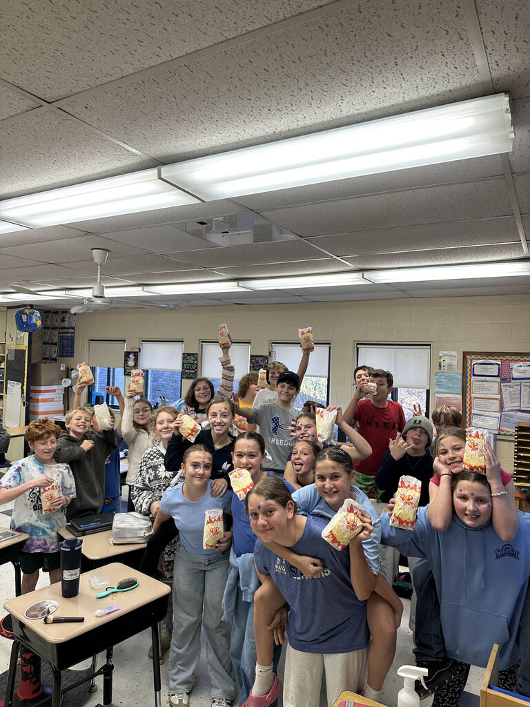 A group of 6th-grade students smiling and posing together in a classroom while holding up individual bags of popcorn.