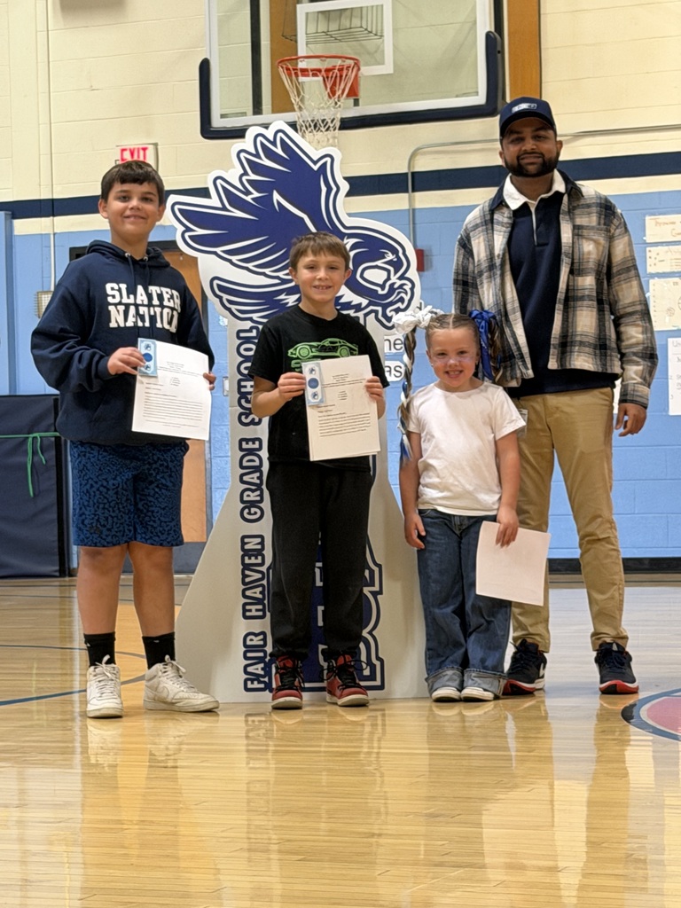 Three elementary students holding certificates pose in the gym with a male teacher in front of the FHGS SOAR standee.