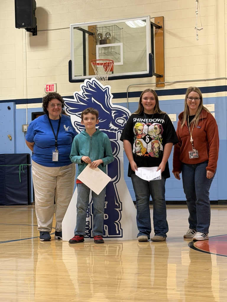 A boy and a girl holding award certificates pose on the gym floor with two teachers next to the SOAR falcon sign.