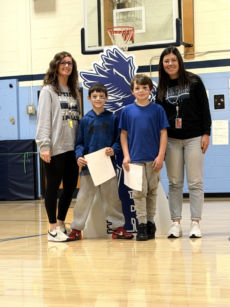 Two boys with blue face paint holding award certificates pose with two teachers beside the FHGS SOAR falcon sign.