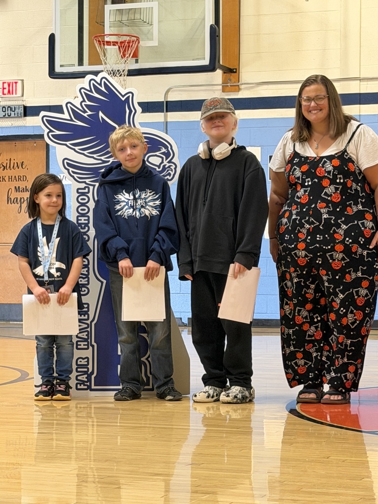 Three students holding award papers pose with a teacher in a skeleton-patterned outfit next to the SOAR falcon cutout.