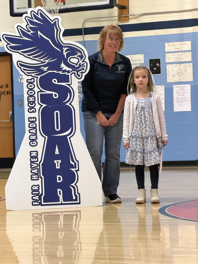 A first-grade student and a teacher standing next to a large blue and white Fair Haven Grade School SOAR falcon cutout on the gym floor.