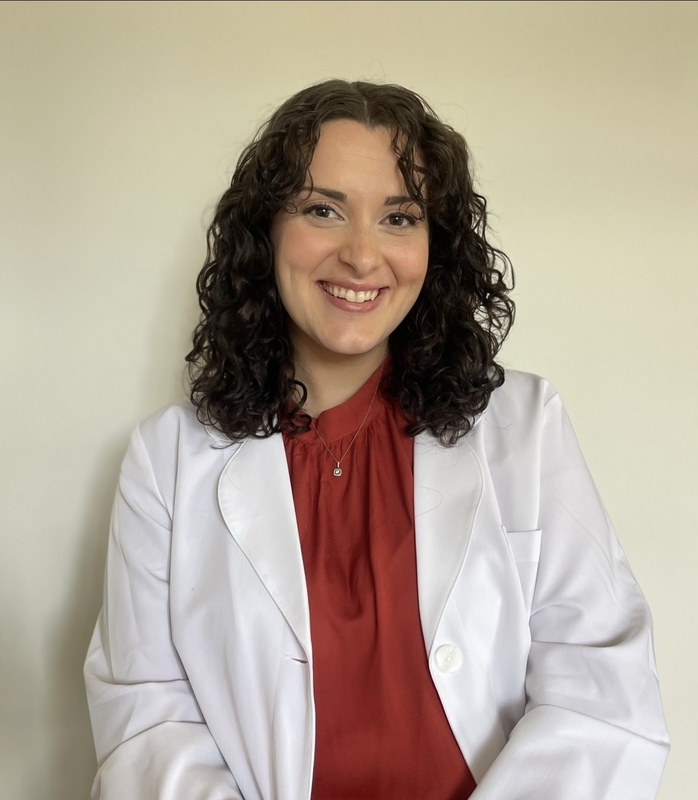 Smiling woman with curly dark hair wearing a white coat over a red blouse, seated against a plain background.