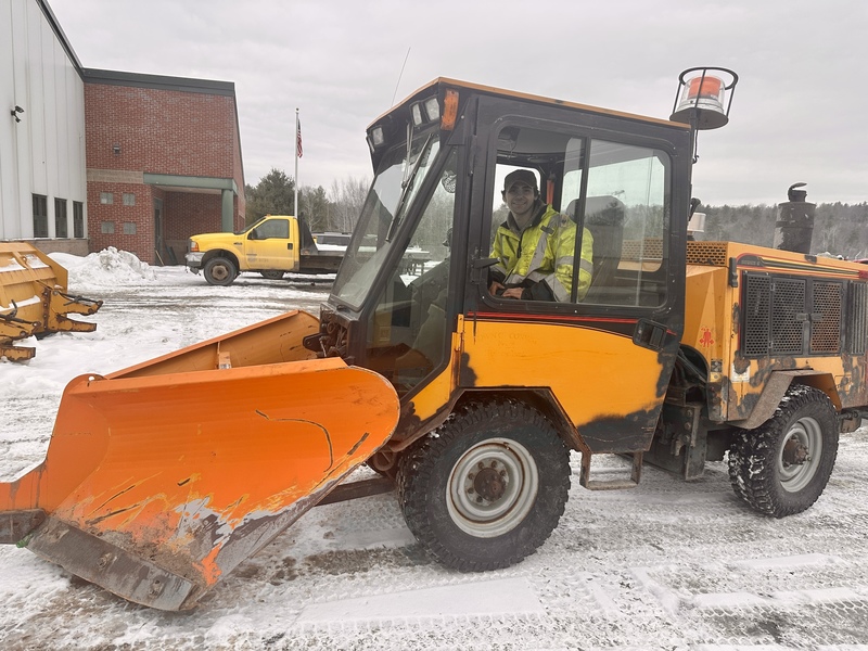 Fair Haven Union High School senior Kaidyn Runnells sits inside a bright orange sidewalk snowplow vehicle at the Fair Haven Public Works Department yard on a winter day. Snow and municipal vehicles are visible in the background near a brick building.
