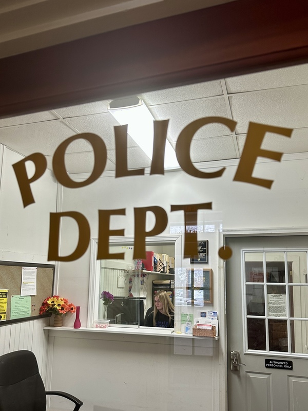 Junior Katie Bouzianis sits at a desk in an office at the Fair Haven Police Department, smiling toward the camera while working at a computer. Office shelves, paperwork, and a bulletin board are visible behind her.