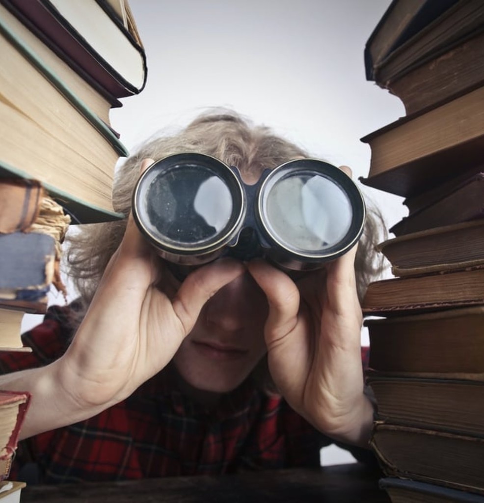 Photo of someone looking through binoculars while standing between two stacks of books