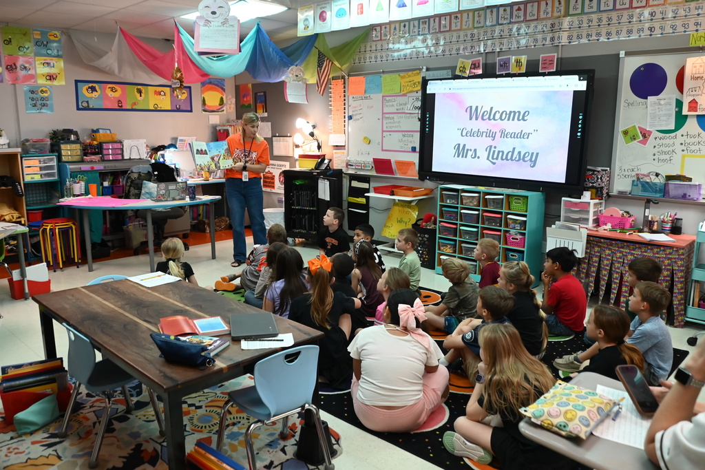 Classroom with a teacher standing in front of a screen, children seated on the floor, chairs, and tables.
