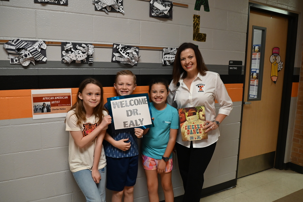 Three children and a woman stand close, smiling, in a hallway. The children hold a sign and a book.