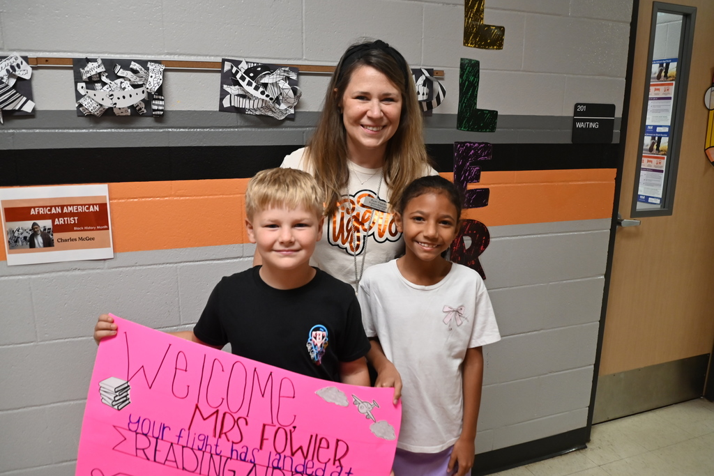 Woman and two children holding a pink sign that says "Welcome Mrs. Fowler". Background includes wall with posters and a door.