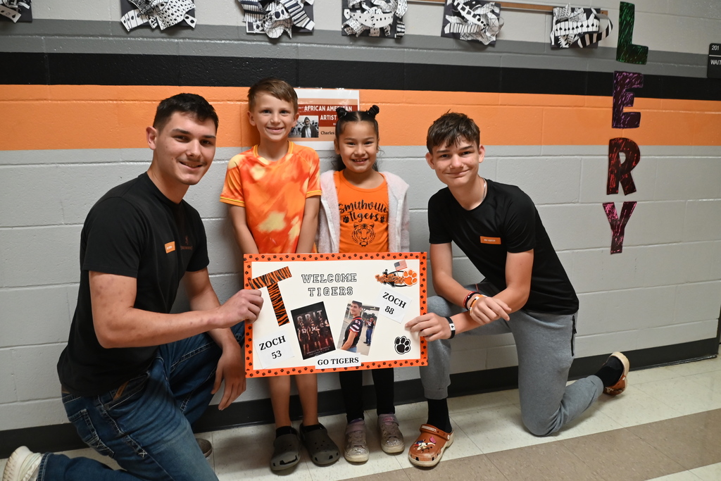 Three people hold a sign that reads "Welcome" against a wall with orange and black stripes.