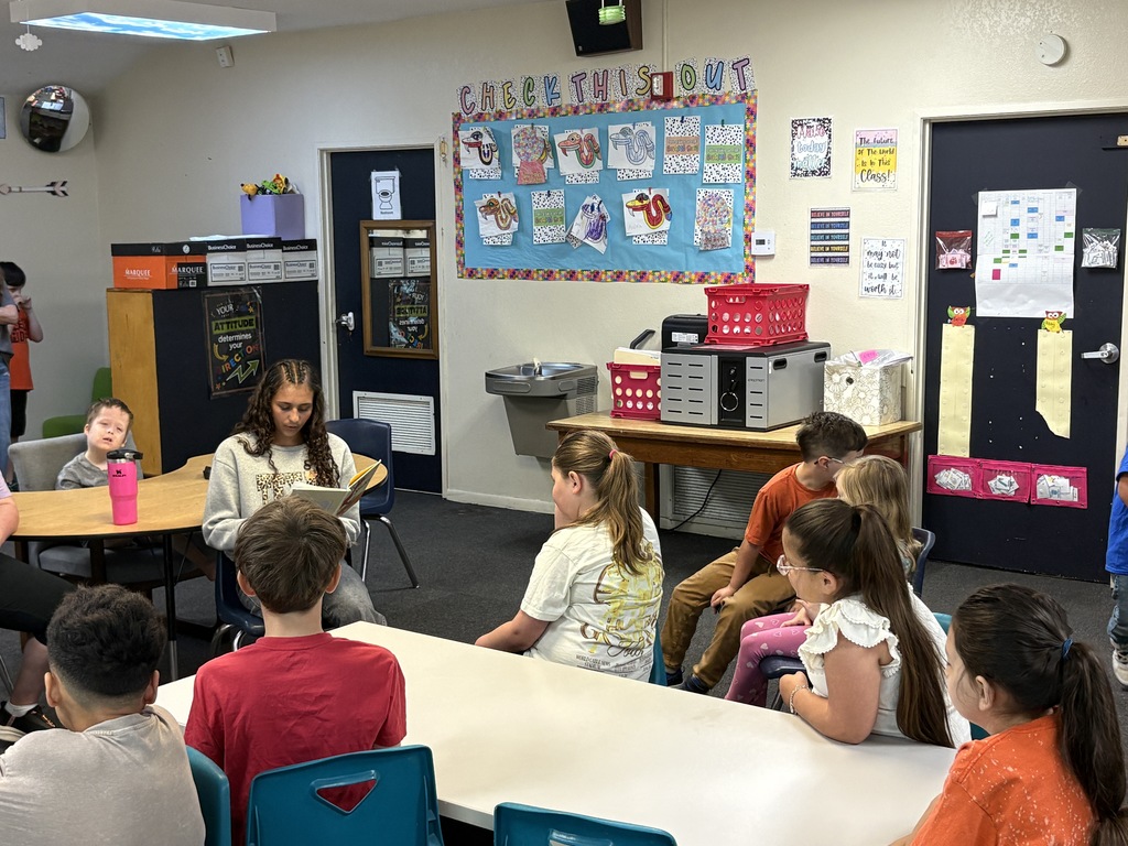 A classroom with tables and chairs, children seated, a woman reading to them, and bulletin boards on the wall.