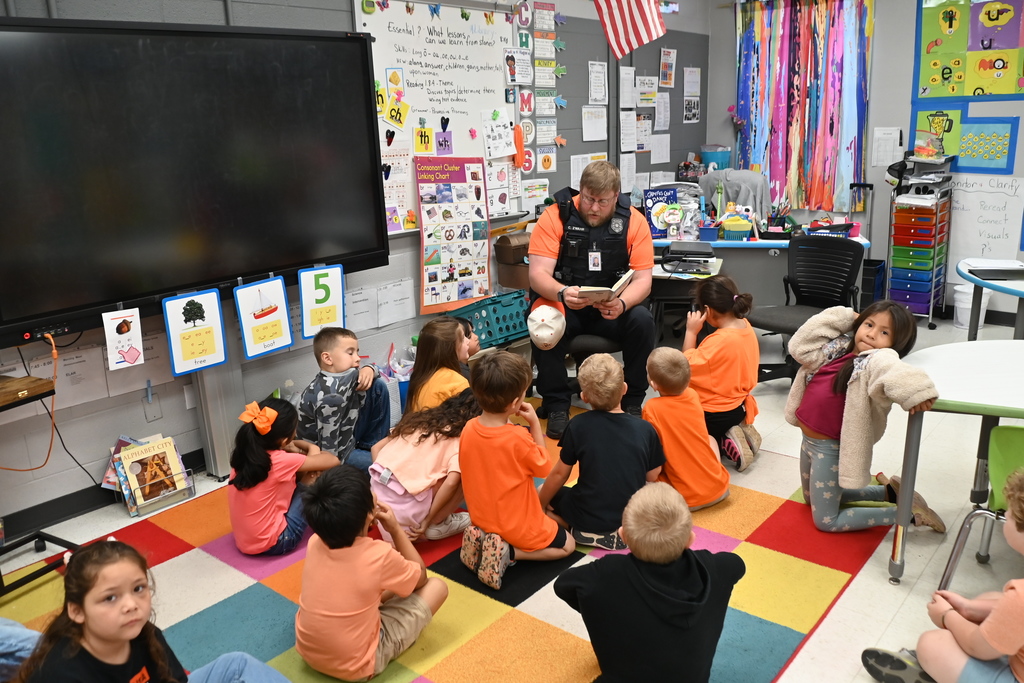 A police officer reads to young children in a colorful classroom with a large monitor and American flag.