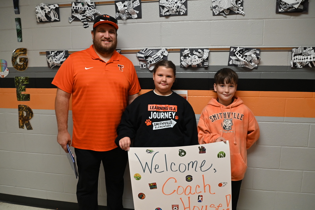 Three people stand in front of a wall with signs. A man wears an orange shirt, and two children wear orange hoodies.