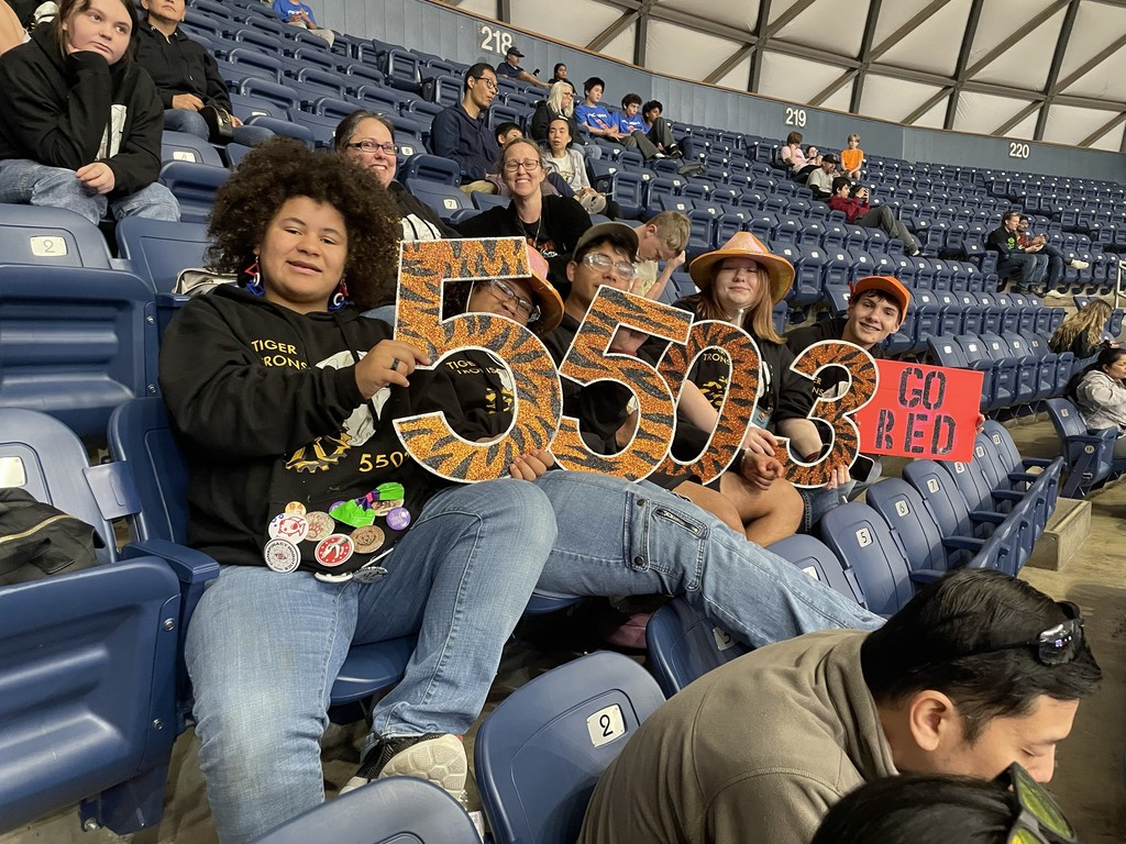 Spectators seated in a stadium. A woman holds up a sign reading 5503. Other fans have hats and signs.