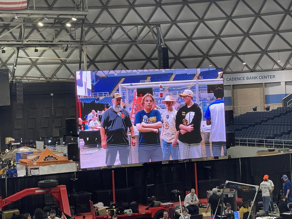 Four people in caps stand on a stage, visible on a large monitor in a large indoor sports venue.