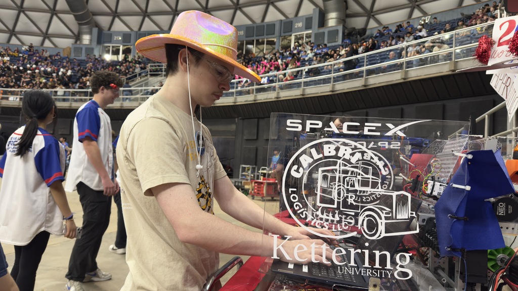 Man in hat working on machine at event. Audience stands behind him. Logo reads "SpaceX Calbri Kettering University".