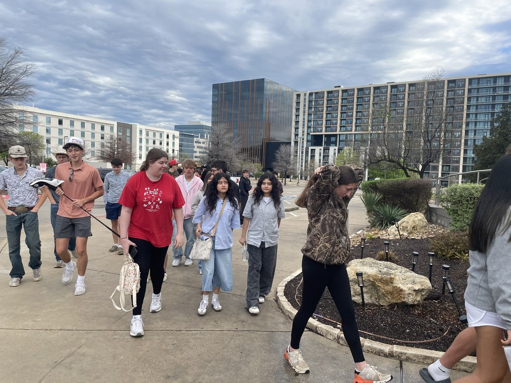 A group of people walk on a sidewalk in front of large buildings. Some wear sneakers and carry bags.