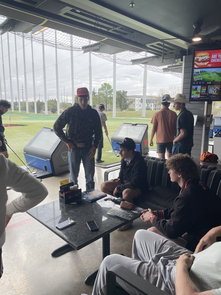 People in a golf lounge, some sitting on a couch and others standing. A large monitor displays a golf game.