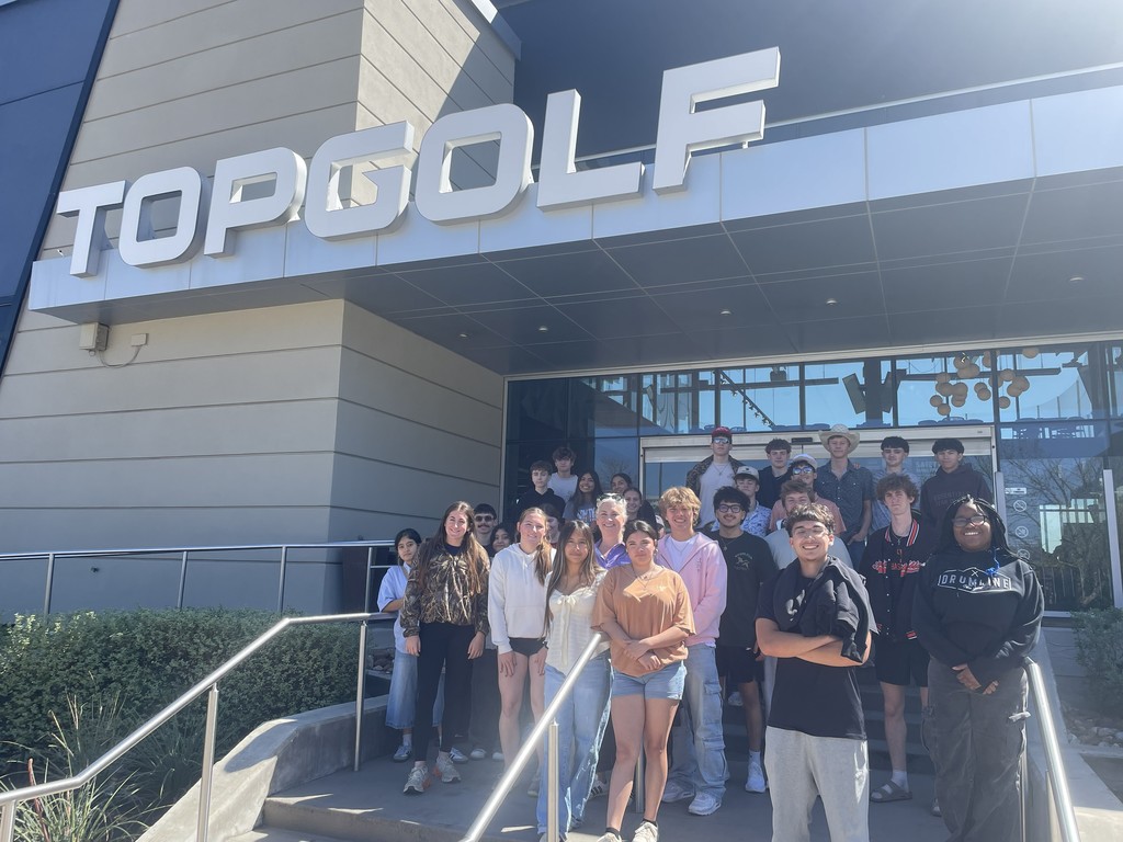 People stand in front of the Top Golf building, with a sign above and steps leading to the entrance.