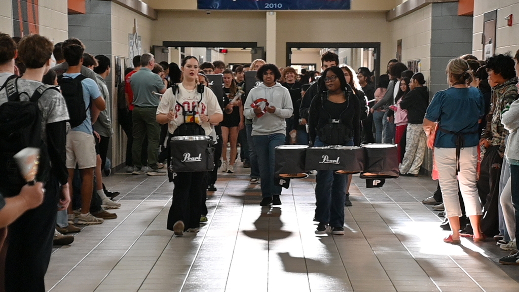 A line of people walking in a hallway carrying drums and other instruments, with onlookers nearby.