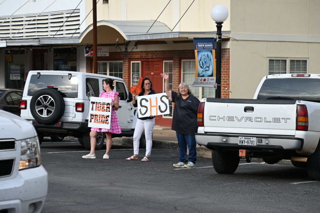 Three people stand in a parking lot with signs. A white SUV is parked behind them, and a building is in the background.