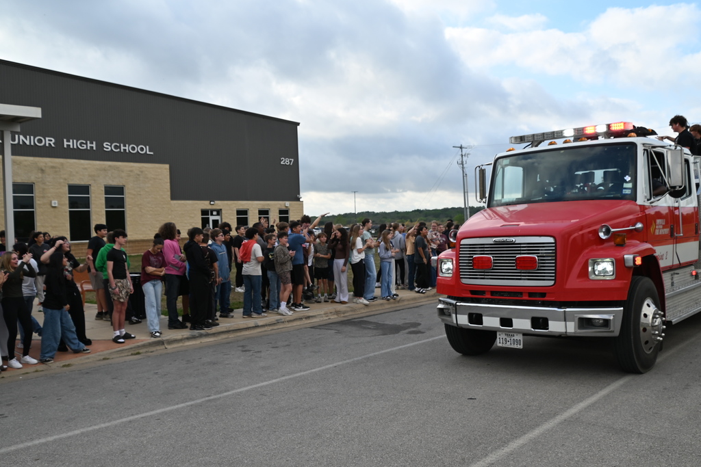 A crowd stands along the sidewalk near a building. A fire truck drives by with flashing lights and a person on the roof.