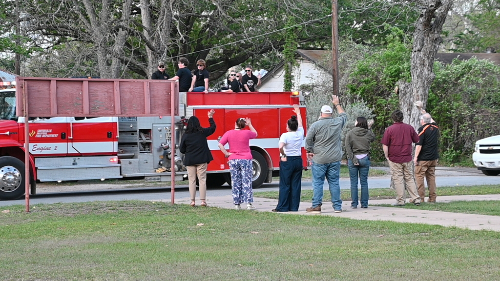 People watch from a lawn as a fire truck drives past with firefighters on the roof.